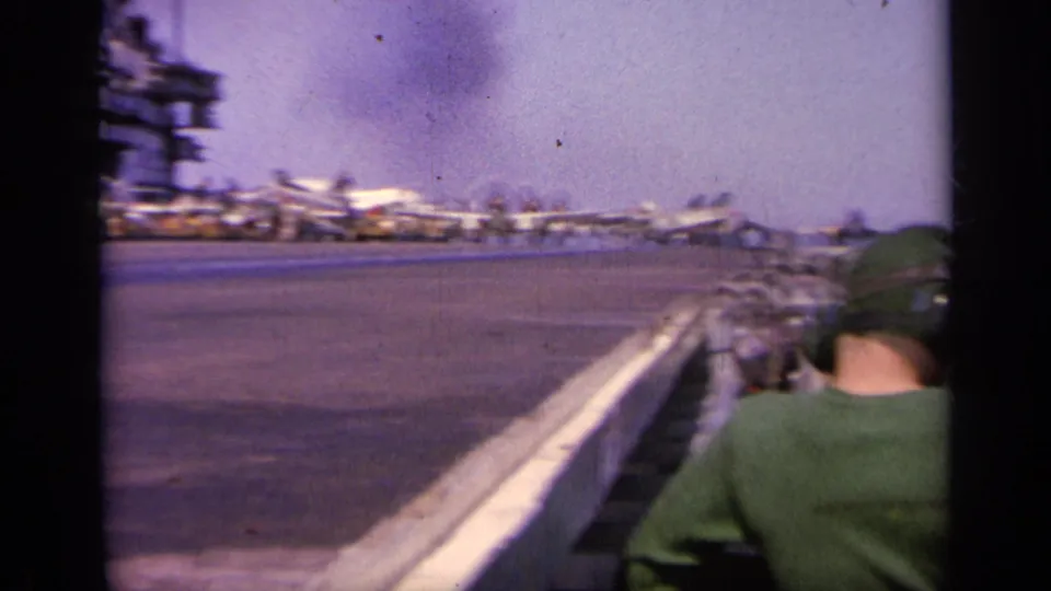 1963 Greece 8mm frame showing a crew member in green near a railing as aircraft sit along the carrier deck.