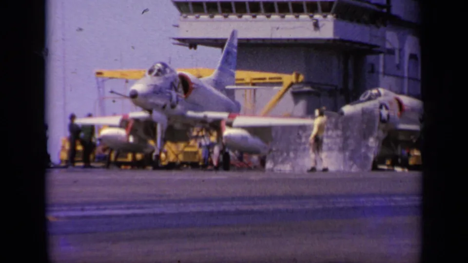 On the carrier deck, crew members stand around a parked military jet near yellow deck equipment and the ship's superstructure.