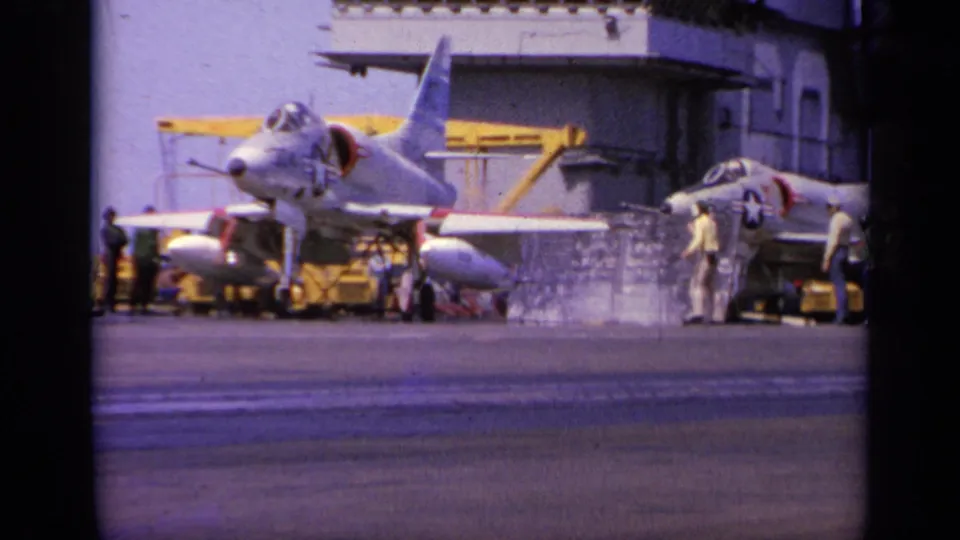 A military jet sits on the carrier deck with crew nearby, yellow equipment behind it, and the carrier island structure filling the background.