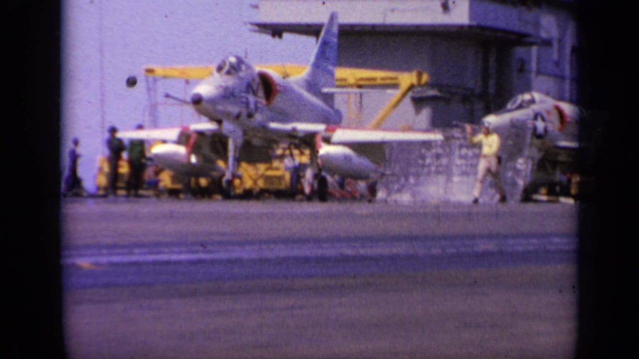 1963 Greece 8mm frame showing a gray U.S. Navy jet on a carrier deck, with crew and deck equipment around it.
