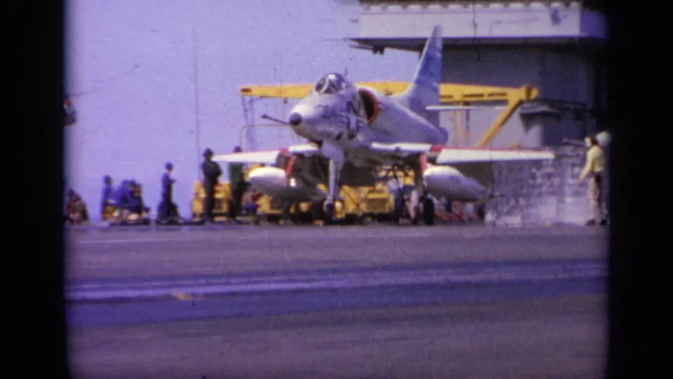 Gray carrier-based jet seen from a distance on a 1963 Greece home-movie frame, with crew gathered near the nose and wing.