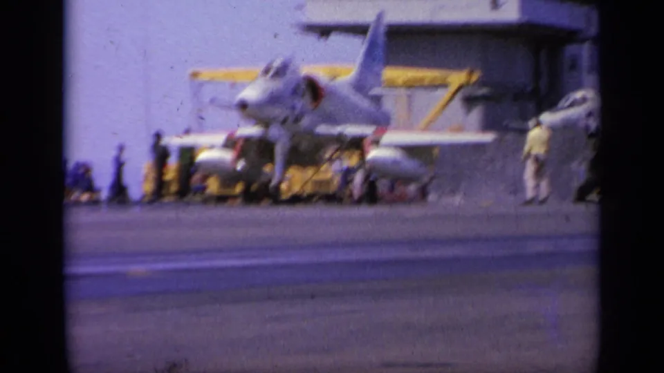 Blurred 8mm frame of a gray naval jet on an aircraft carrier deck in the 1963 Greece clip, with crew and support gear visible.