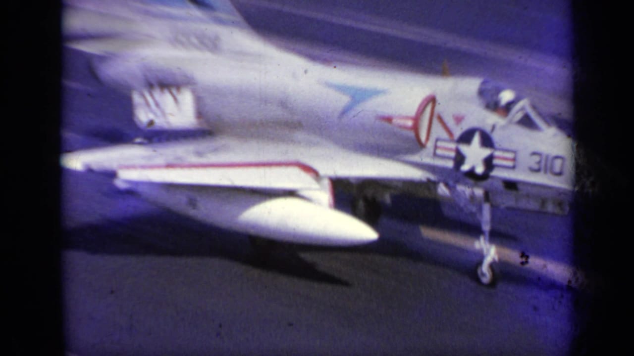 1963 8mm frame showing a U.S. Navy-marked jet near the carrier deck with ocean visible beyond and dark film gate edges.