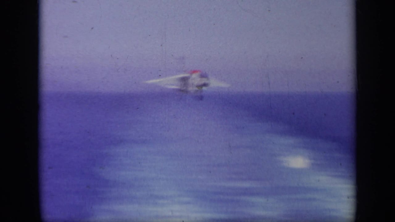 Jet approaching over the ocean toward an aircraft carrier.
