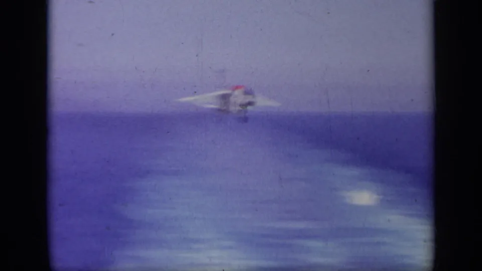 1963 8mm frame showing a Navy jet approaching over open water, seen from the carrier area with soft focus and color shift.