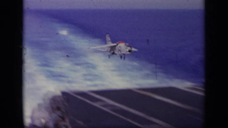 1963 8mm frame showing a Navy jet descending toward the carrier deck, with ocean wake or water texture behind it.