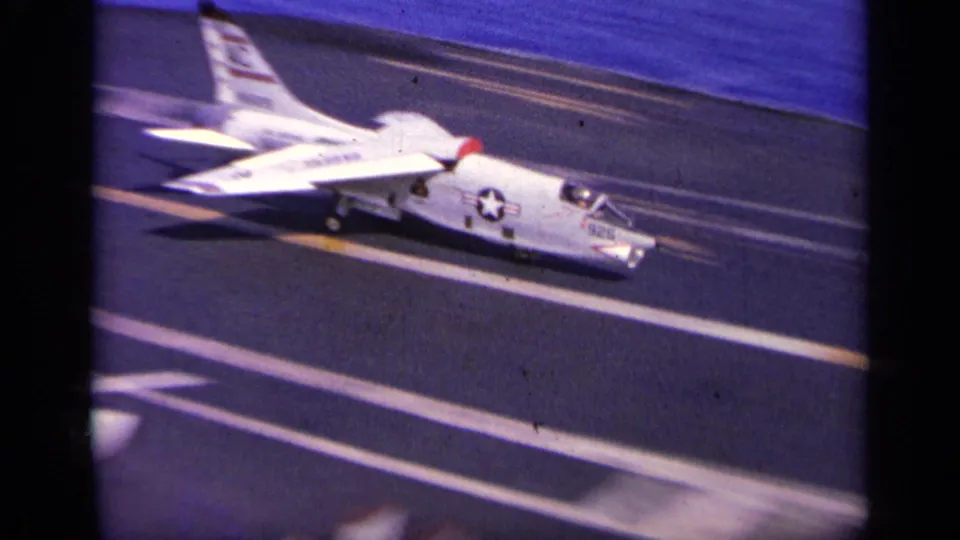 1963 8mm frame showing a U.S. Navy jet on the carrier flight deck, with deck striping, insignia, and aircraft markings visible.