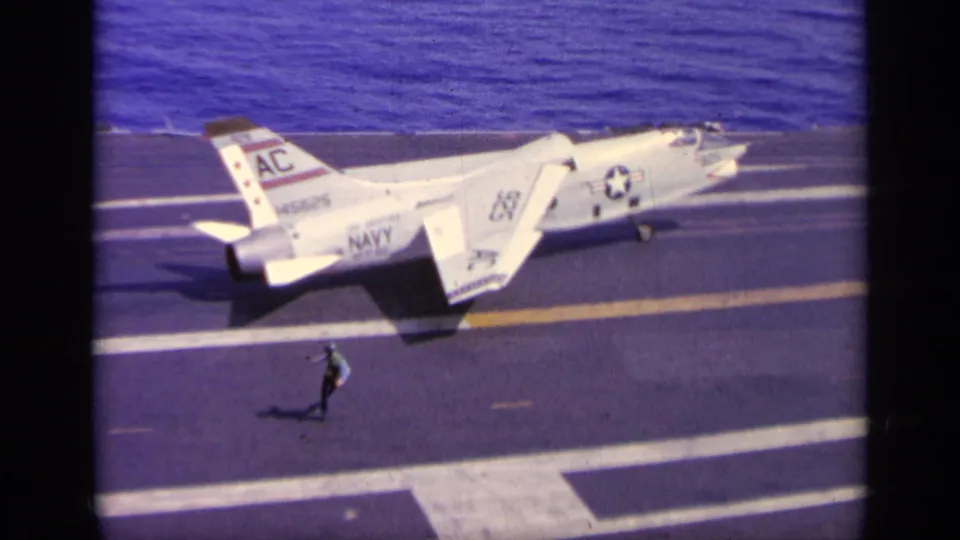 A white U.S. Navy jet taxis on a carrier deck while a crew member moves across the foreground in 1963 Greece footage.