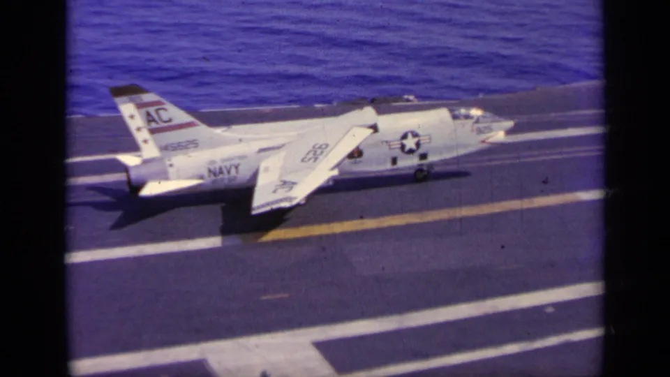 Side view of a white U.S. Navy aircraft with AC tail marking taxiing on an aircraft carrier deck near the sea.