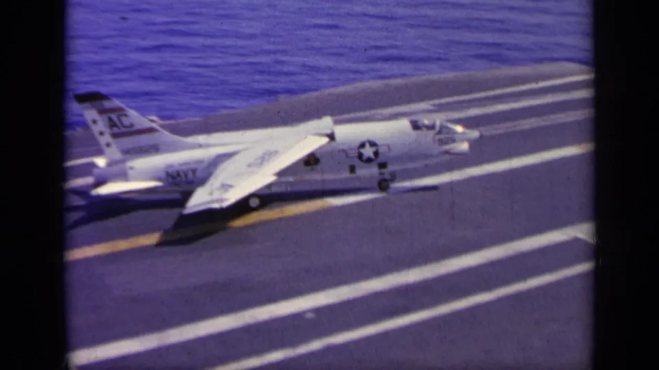 The Navy jet continues taxiing across the carrier deck, framed by runway markings and open water in the background.