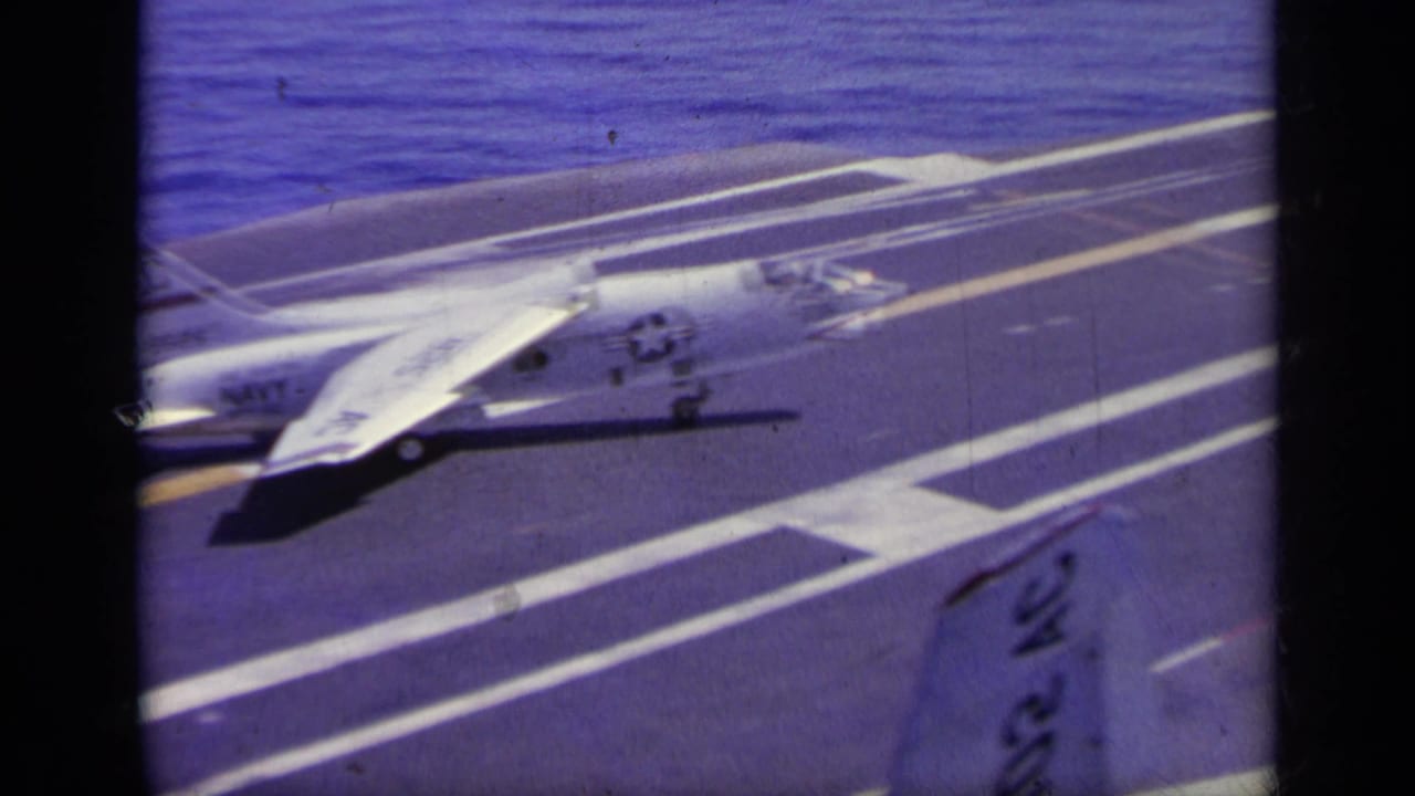 1963 Greece metadata supports this 8mm view of a white naval jet on a carrier deck with runway markings and sea in the background.