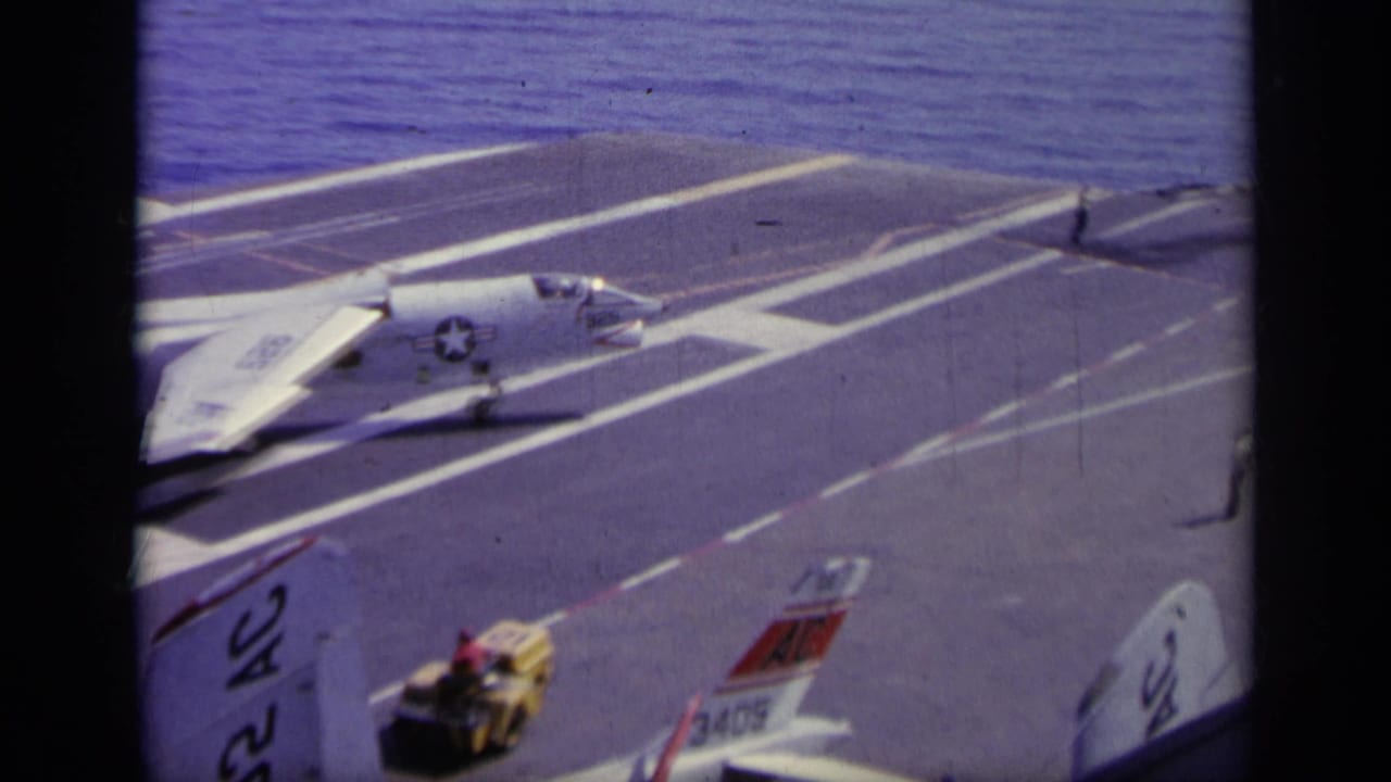 Naval jet on a carrier runway with deck equipment and aircraft tails nearby.
