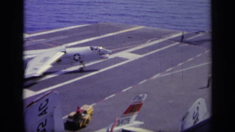 White military jet appears on the carrier flight deck, with marked runway lines, nearby deck vehicle, aircraft tails, and open sea visible.