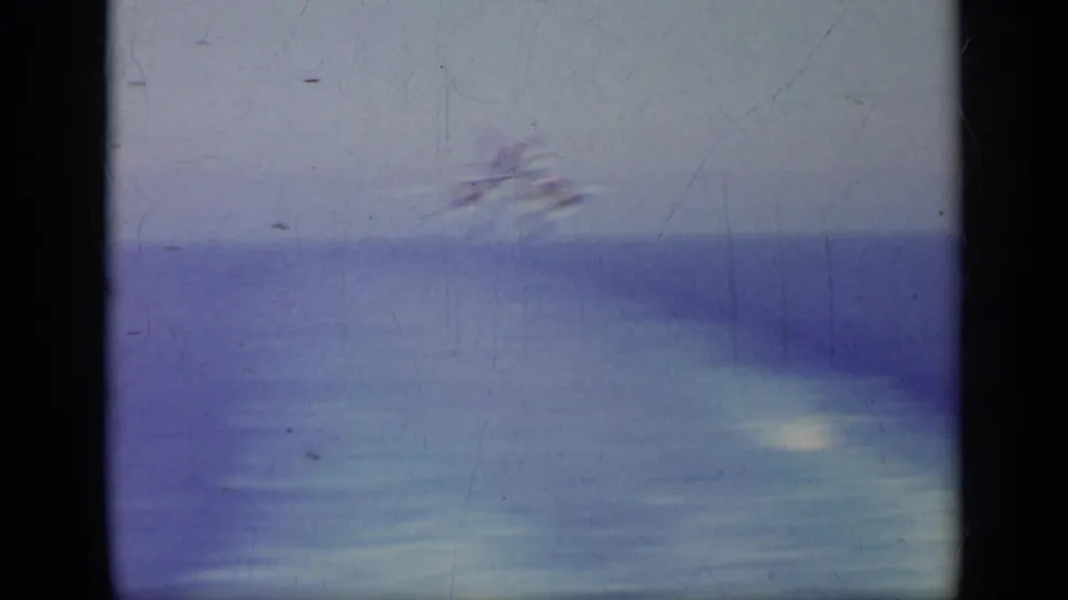 A blurred 8mm frame shows bright haze or exhaust over the carrier deck, likely during aircraft movement, with the sea-toned background visible.