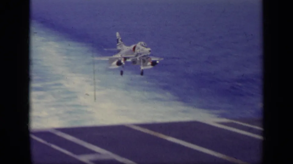 A white naval jet appears just above or near the aircraft carrier deck, with exhaust or spray behind it and deck runway markings below.