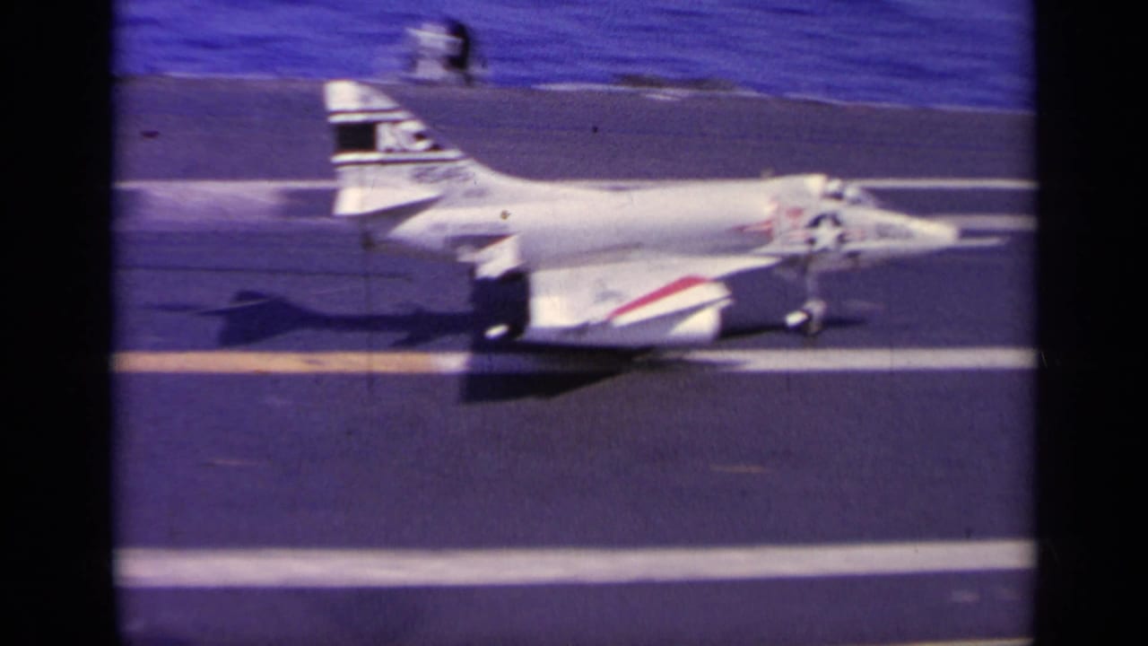 Navy jet rolls along a carrier deck with ocean in the background.