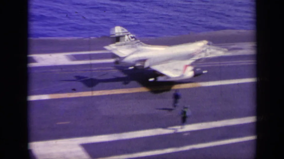 1963 8mm frame showing the Navy jet moving past visible deck crew on an aircraft carrier deck at sea.