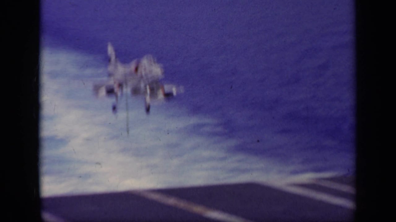 1963 Greece 8mm frame showing a white naval jet approaching an aircraft carrier deck with ocean behind it.