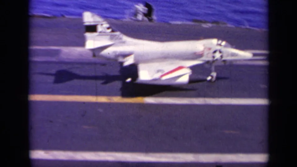 Side view of the white naval jet rolling on the aircraft carrier deck, with tail markings and red-blue wing striping visible.