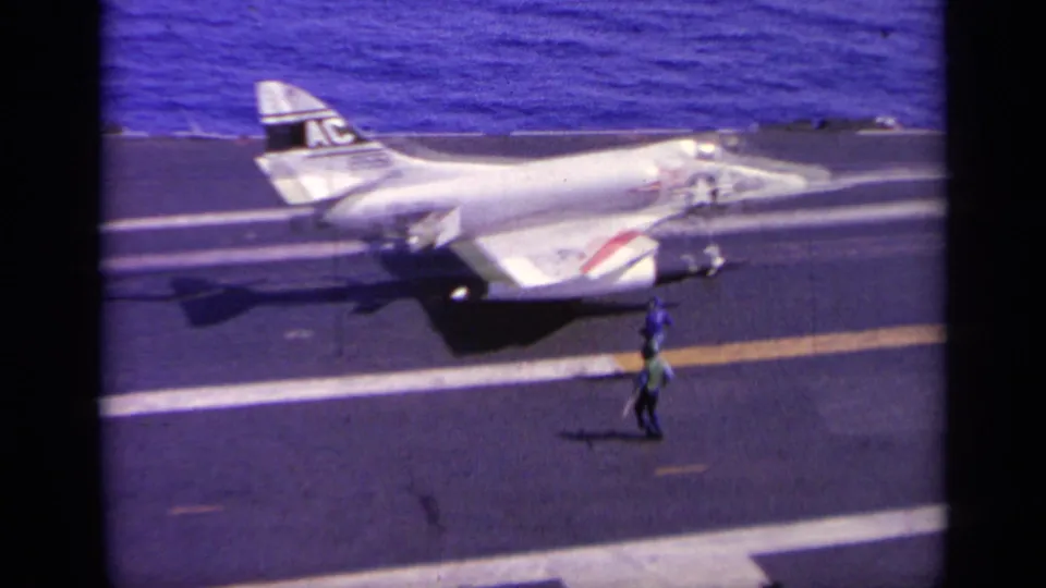 White naval jet on the carrier deck in 1963 Greece footage, with ocean behind it and a deck crew figure walking nearby.