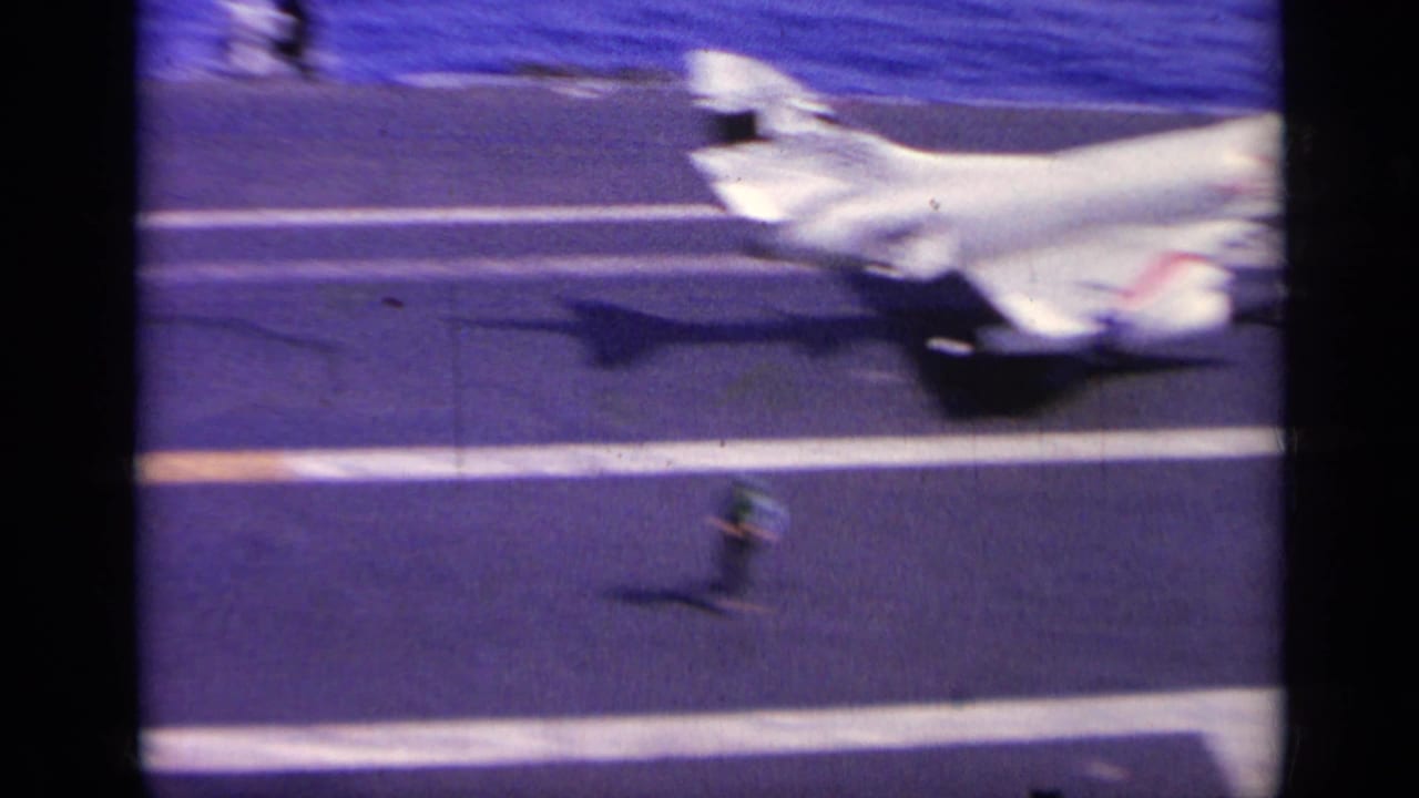 1963 Greece home-movie frame showing a white military jet with red markings on an aircraft carrier deck at sea.