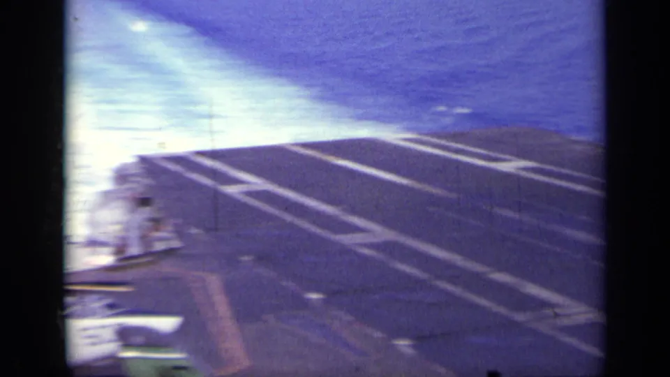Carrier deck at sea with bright wake, deck markings, and small crew figures visible near the ship edge.