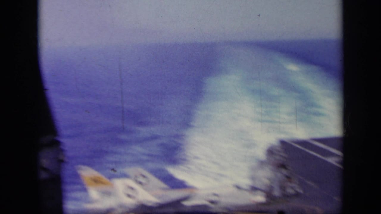 1963 Greece 8mm frame showing a military jet near an aircraft carrier deck, with sea horizon, motion blur, and visible film gate edges.
