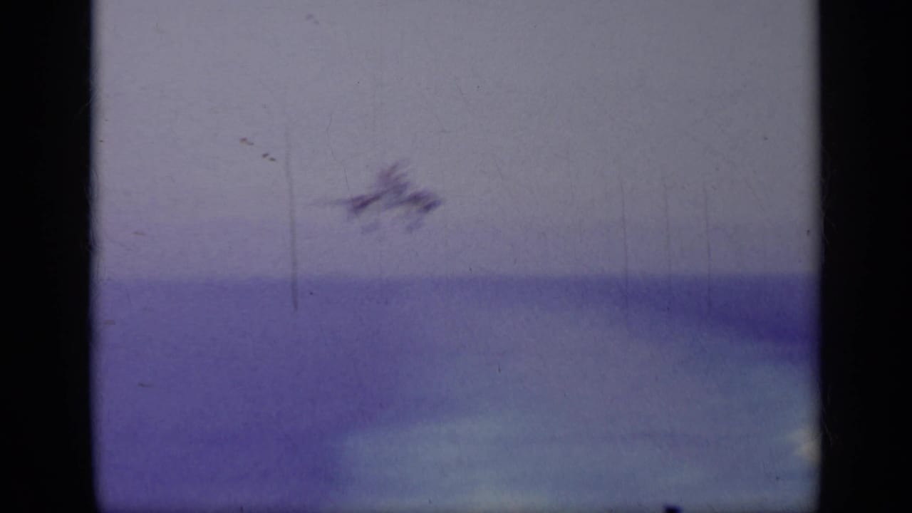 Small blurred aircraft over ocean with dark vertical film gate bars.