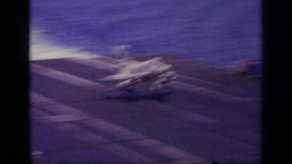 1963 Greece 8mm frame showing a military jet on a carrier flight deck at sea, with white deck markings and a hazy horizon.