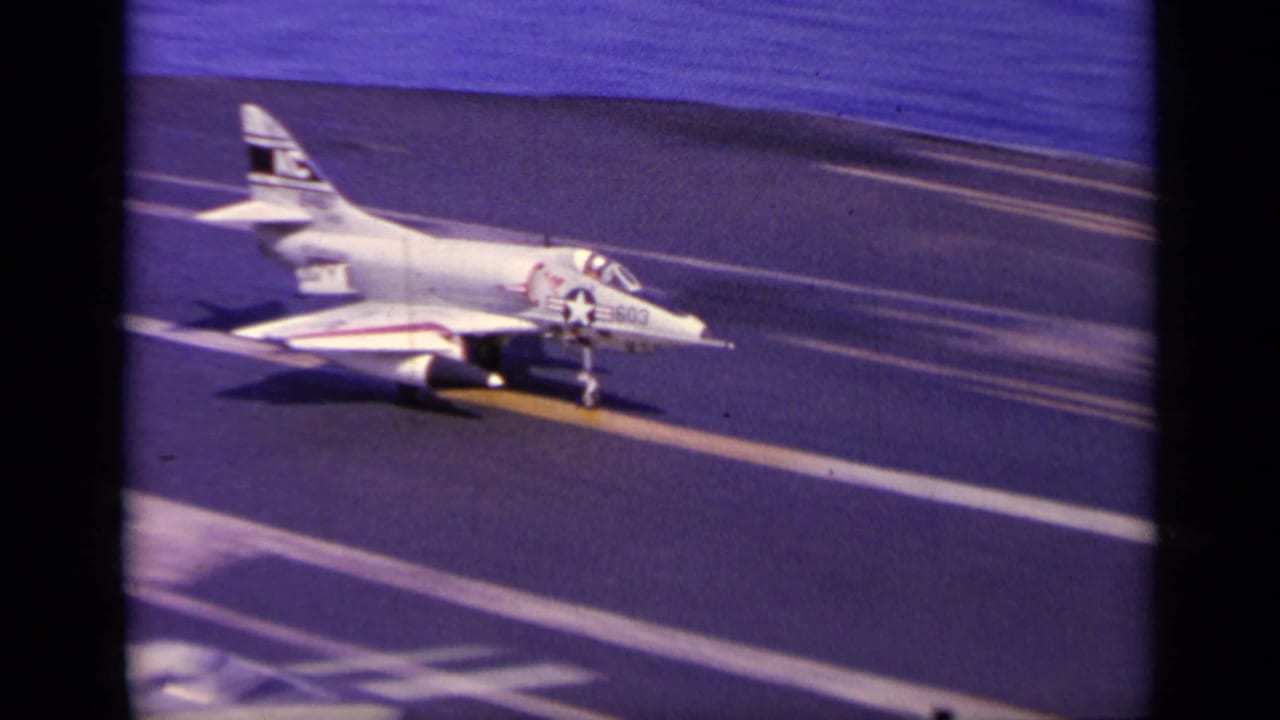 1963 8mm frame of a white U.S. Navy jet approaching a carrier deck near Greece, with tail markings and sea visible behind it.