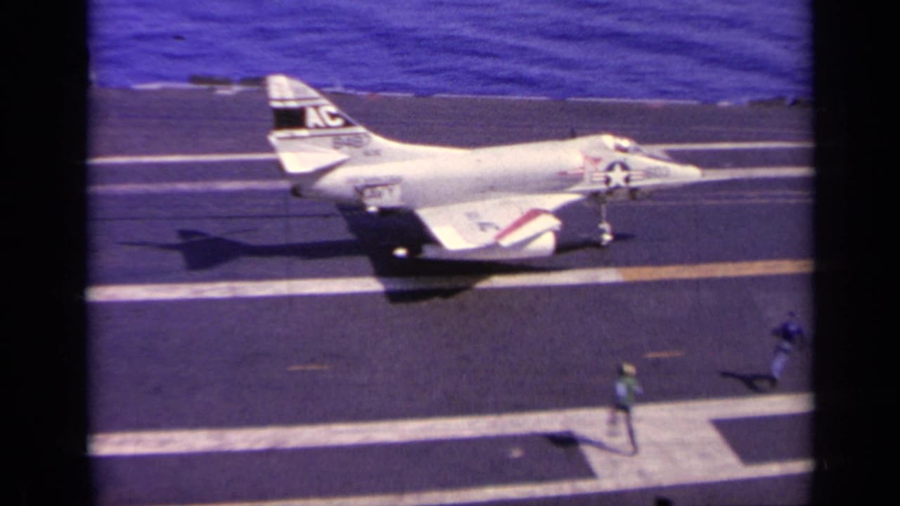 White Navy jet rolling on the aircraft carrier deck with small crew figures nearby.