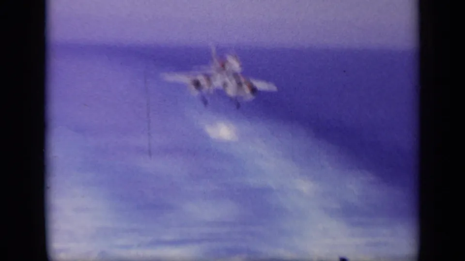 Blurred 1963 8mm frame showing the Navy jet in motion over the carrier deck area, with blue ocean and film softness dominating the view.
