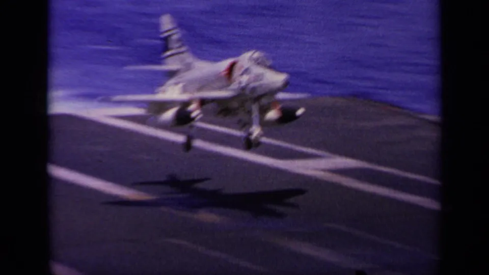 A white Navy jet sits or rolls on the carrier deck in 1963 Greece footage, with a dark aircraft shadow and ocean visible behind it.