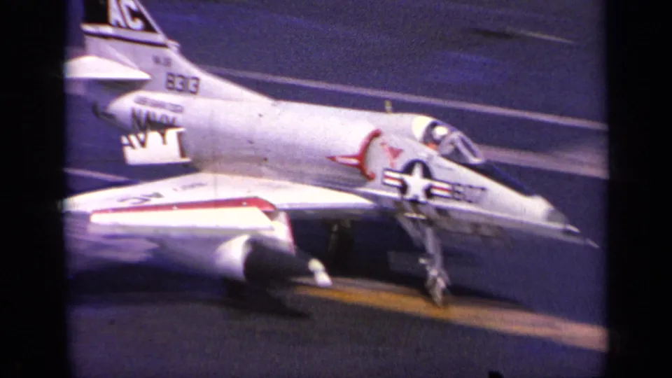 1963 Greece home-movie frame of a U.S. Navy jet landing on an aircraft carrier, with tail code and deck markings visible.