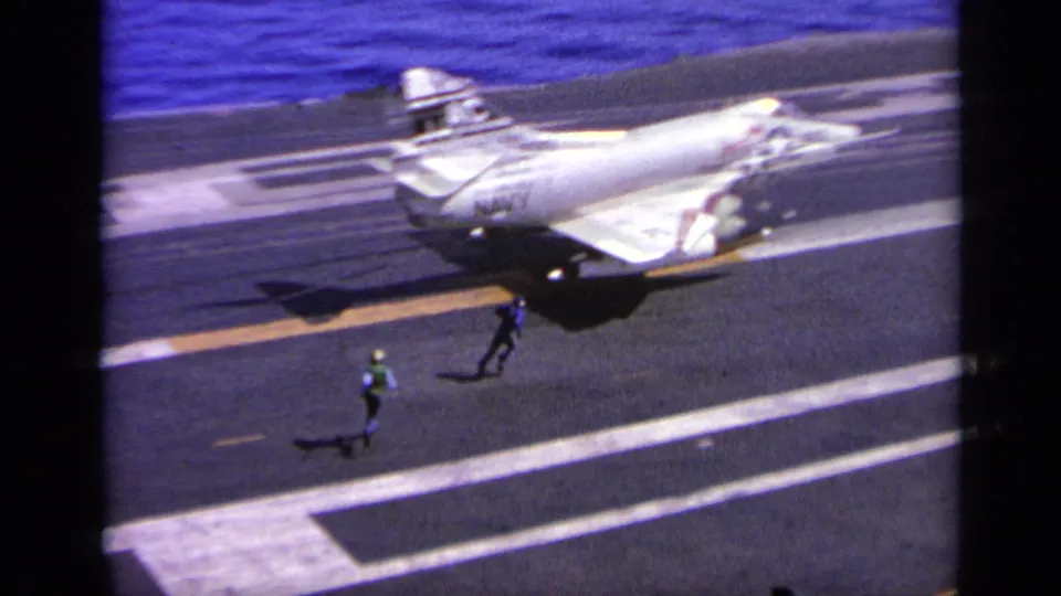 The Navy jet decelerates on the carrier flight deck as a deck crew member in a bright vest stands below the aircraft.