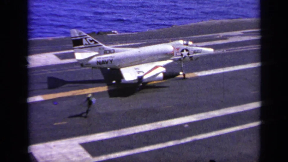 A U.S. Navy jet rests or slows on the carrier deck, with a crew member crossing in the foreground and ocean beyond.