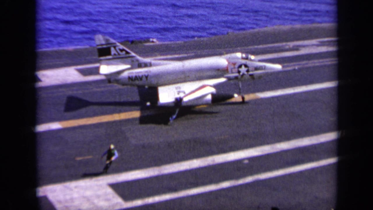 1963 Greece home-movie frame showing a U.S. Navy-marked jet on a carrier deck as a crew member stands nearby.