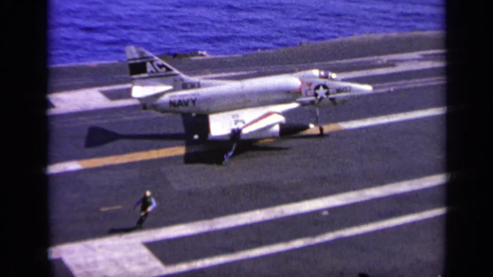1963 Greece home-movie frame showing a U.S. Navy-marked jet on a carrier deck as a crew member stands nearby.