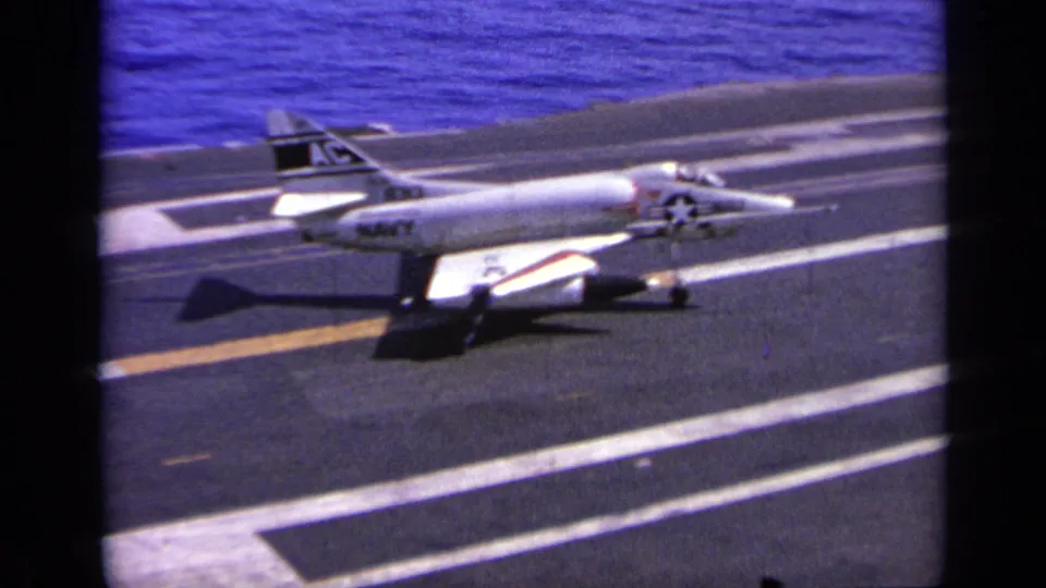A 1960s U.S. Navy-marked jet appears on the aircraft carrier deck, with flight markings and blue sea in the background.