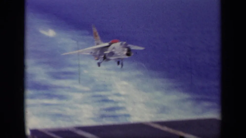 The Navy jet is airborne above the carrier deck in a 1963 Greece home-movie view, with sky, sea, and runway markings below.
