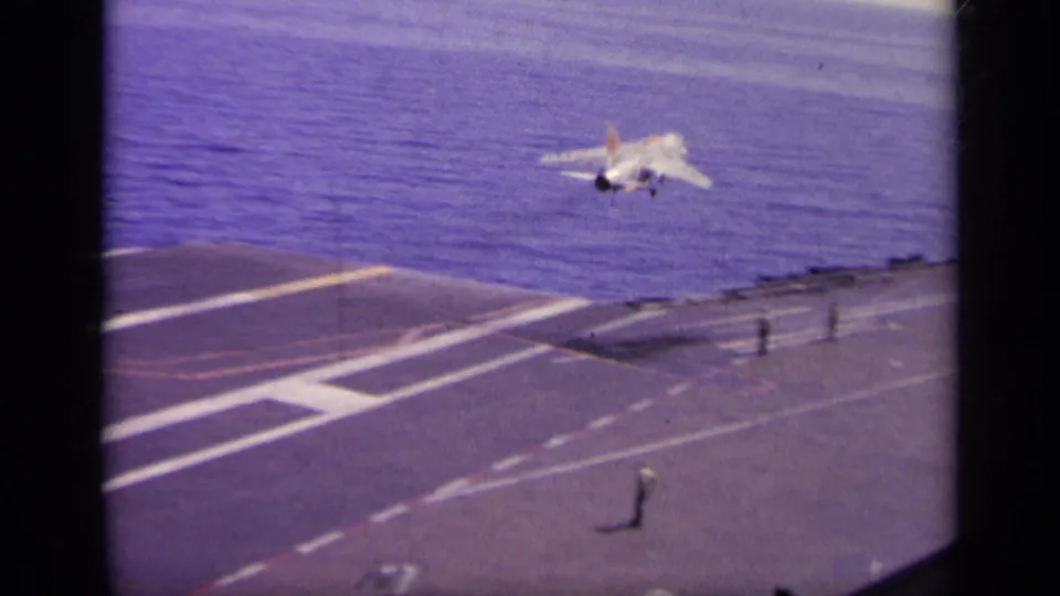 The Navy jet rises from the aircraft carrier deck, with runway striping, deck fixtures, and open water behind it.