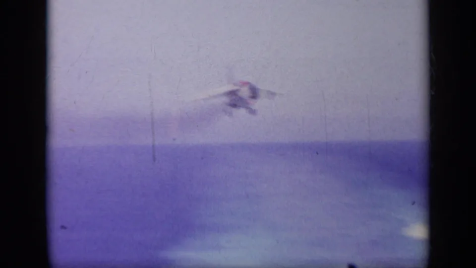 Soft, grainy 8mm frame of the Navy jet airborne over blue water after leaving the carrier deck.