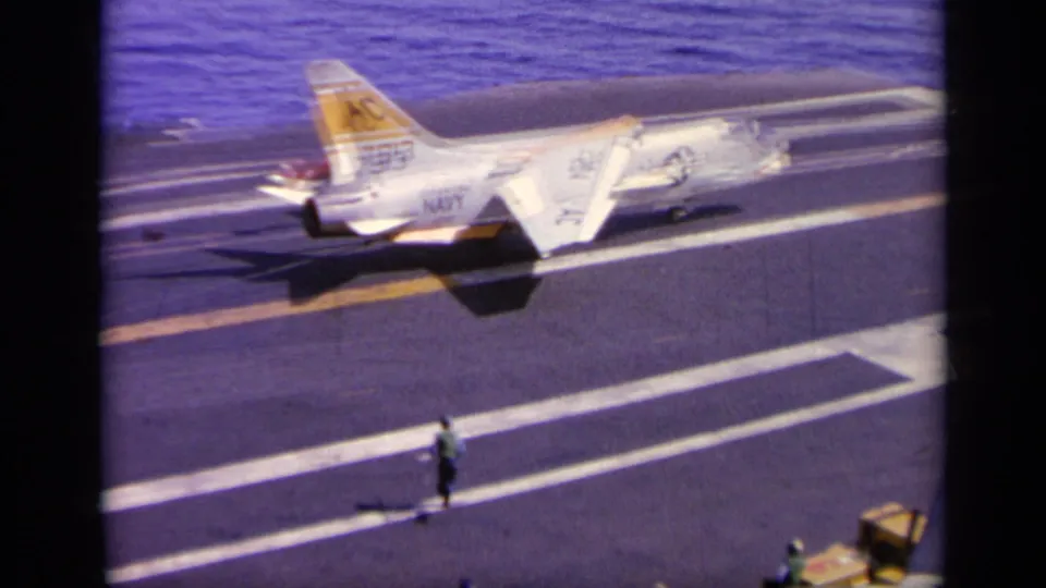 1963 8mm frame showing a U.S. Navy jet on the carrier deck as crew members stand near the marked landing area.