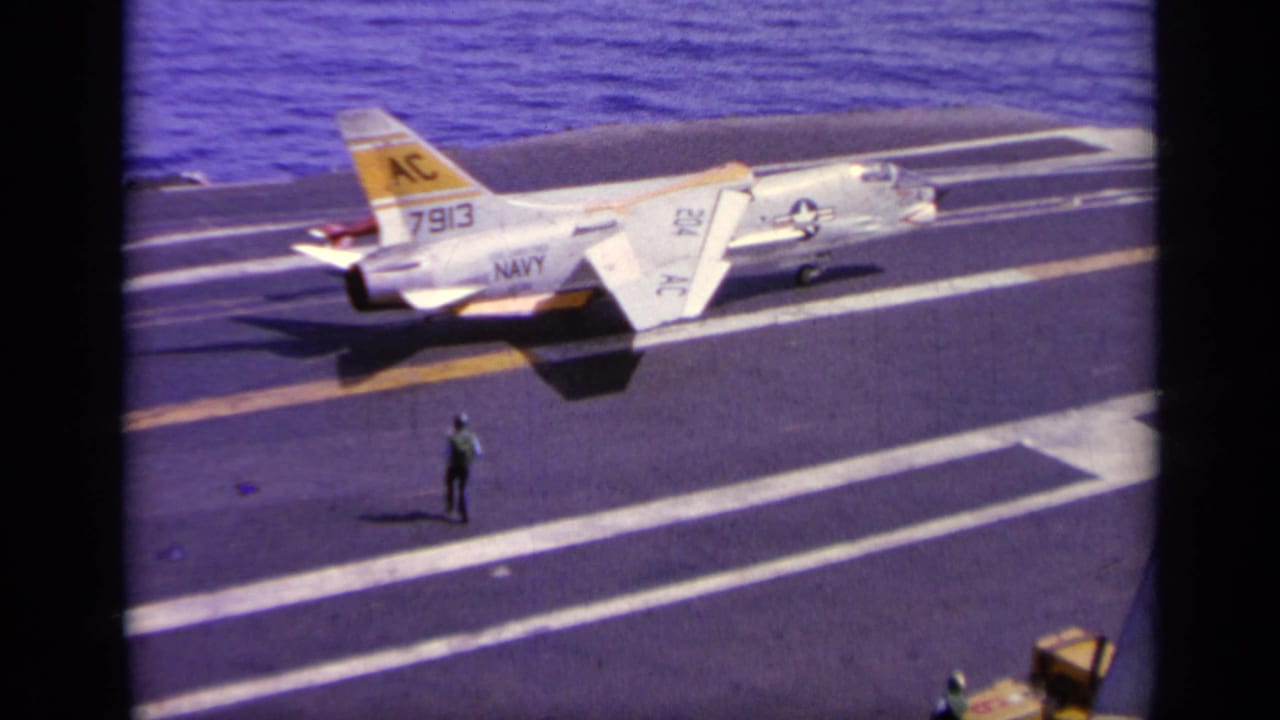 1963 8mm footage near Greece showing a marked U.S. Navy jet on an aircraft carrier deck with ocean in the background.