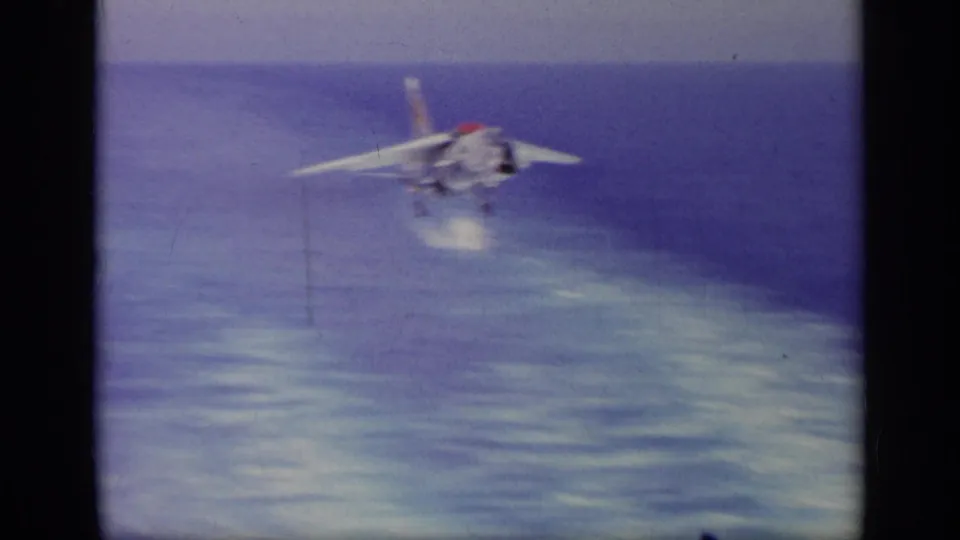 A Navy jet appears over the sea on approach, with landing gear visible and the horizon behind it.