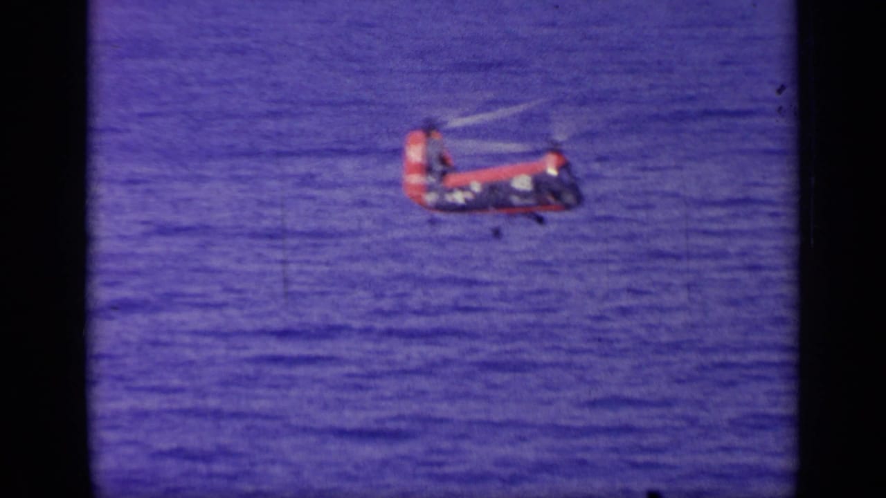 Small red and blue helicopter flying above the ocean.