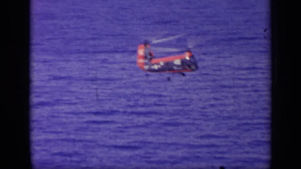 Small red and blue helicopter hovers or flies low over deep blue water in 1963 Greece-associated home-movie footage.