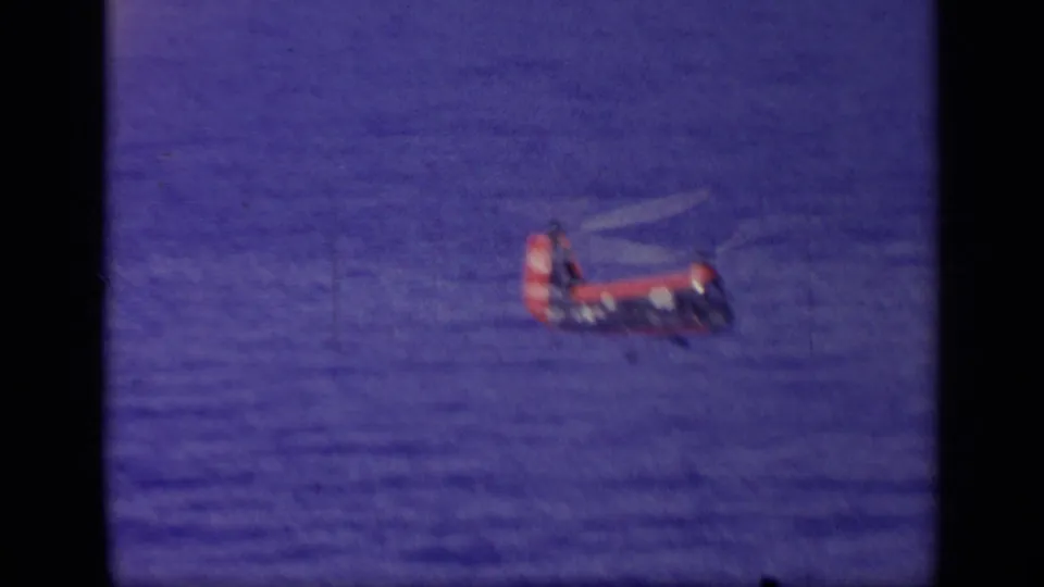 Distant red and blue helicopter remains over the ocean, softened by grain and motion in the 8mm frame.