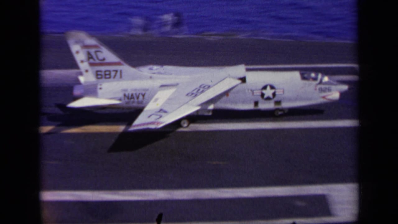 1963 home-movie frame from Greece showing a white U.S. Navy jet marked AC 6871 on a carrier flight deck, with blue sea beyond.