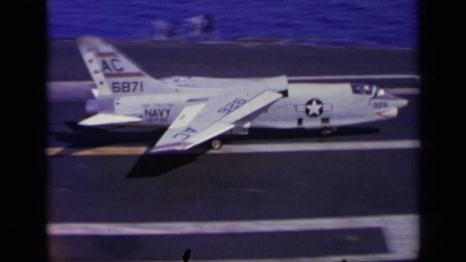 1963 home-movie frame from Greece showing a white U.S. Navy jet marked AC 6871 on a carrier flight deck, with blue sea beyond.